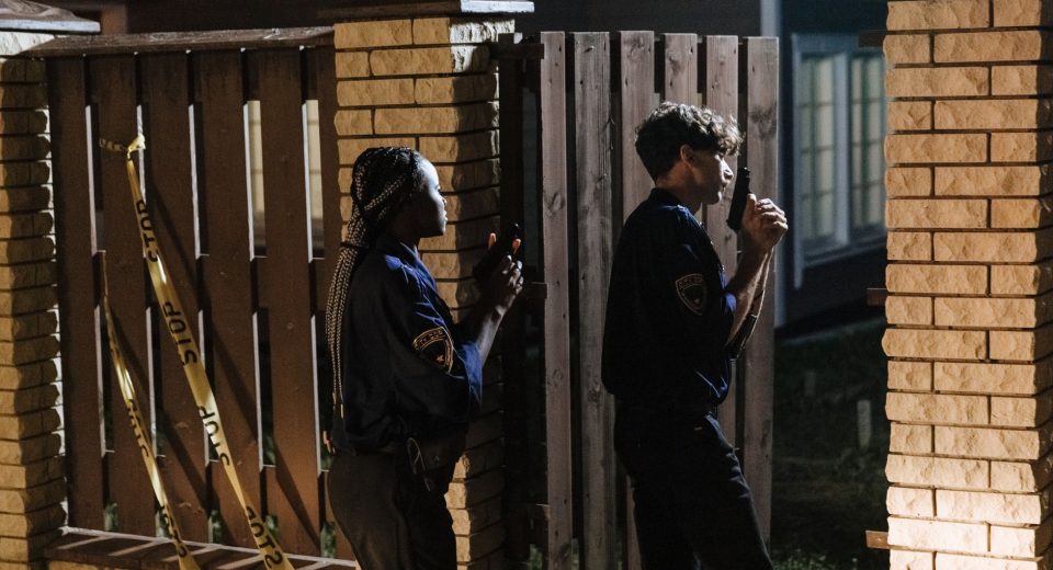 Dois policiais entram com cautela em uma residência durante uma investigação noturna.