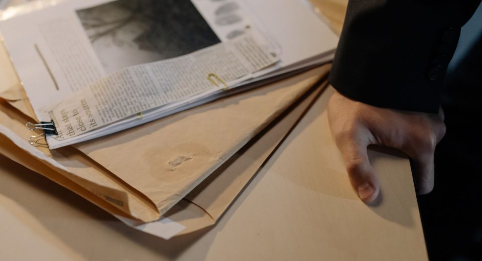 Mão apoiada sobre uma mesa ao lado de documentos de investigação e recortes de jornal.
