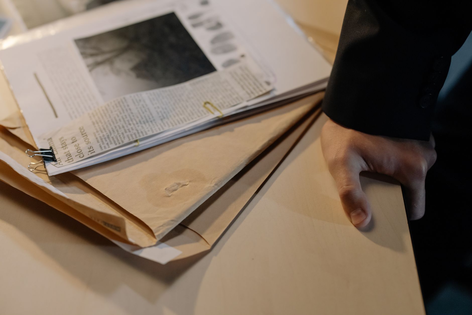 Mão apoiada sobre uma mesa ao lado de documentos de investigação e recortes de jornal.