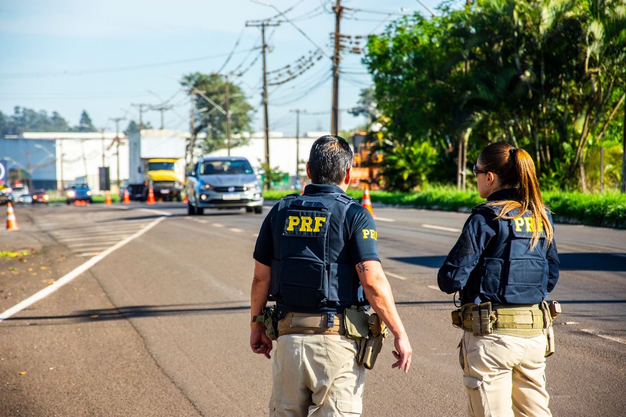 Agentes da Polícia Rodoviária Federal monitoram o trânsito em uma rodovia brasileira sob céu ensolarado.