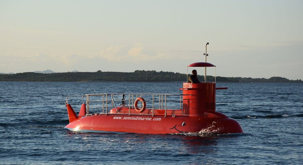 Embarcação semissubmersível vermelha navegando em águas calmas, com uma faixa de terra ao fundo