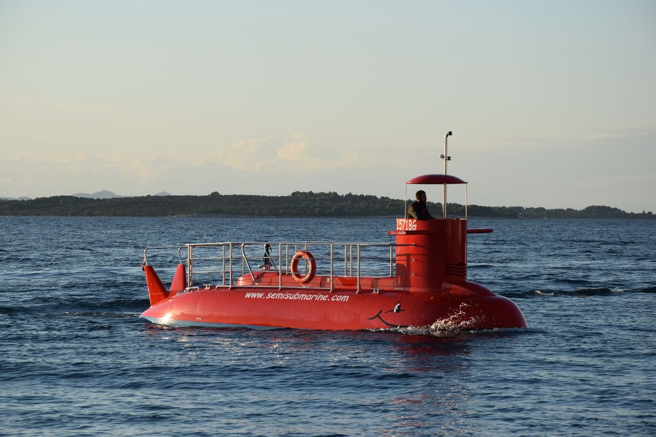 Embarcação semissubmersível vermelha navegando em águas calmas, com uma faixa de terra ao fundo