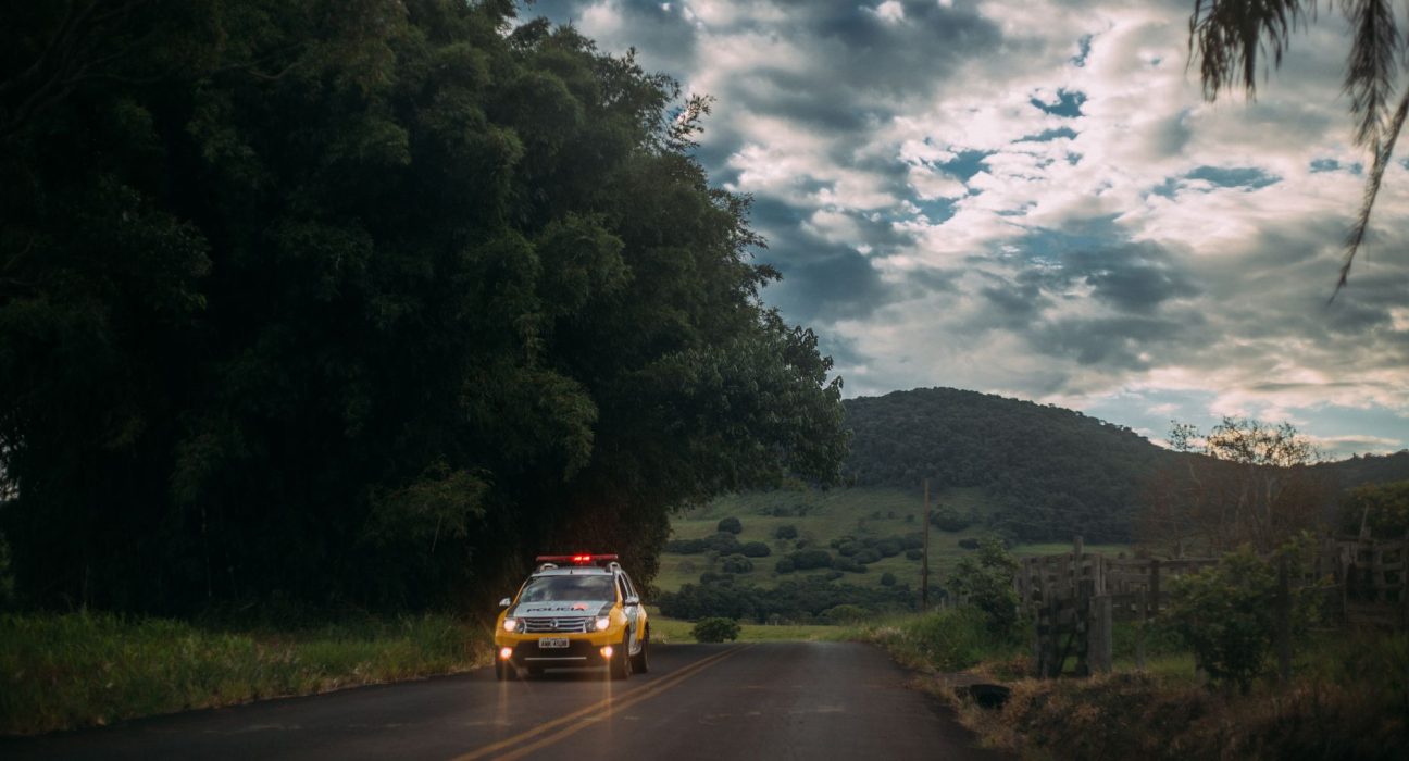 Viatura policial percorre uma estrada rural ao entardecer.