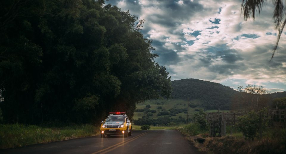 Viatura policial percorre uma estrada rural ao entardecer.