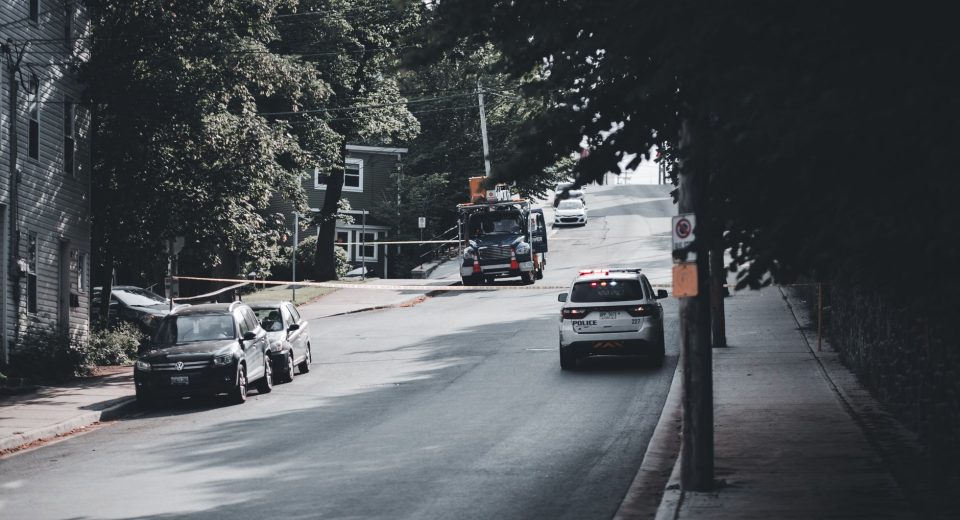 Viatura policial circula por uma rua urbana arborizada, com carros estacionados nas laterais.