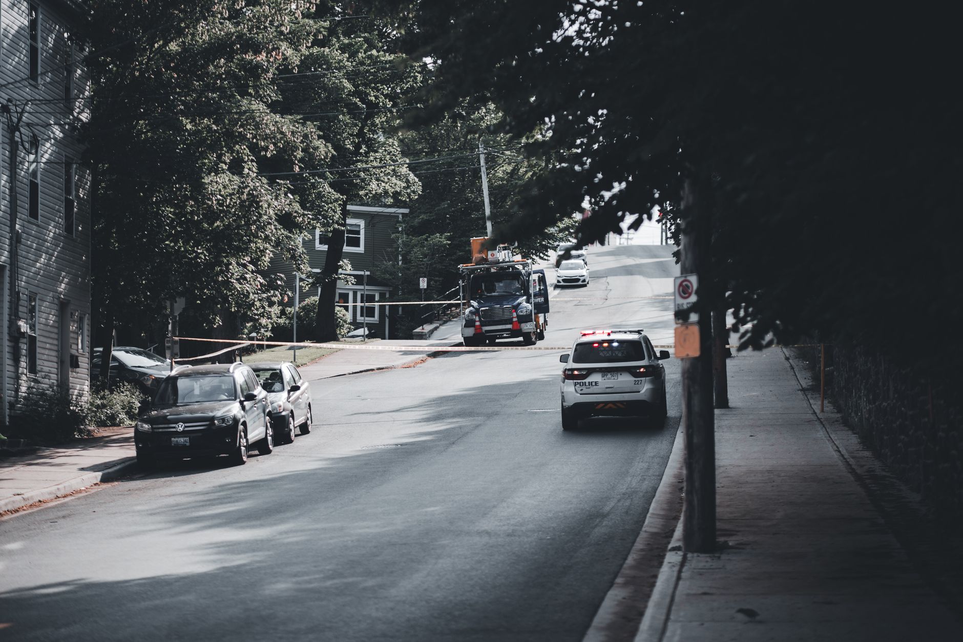 Viatura policial circula por uma rua urbana arborizada, com carros estacionados nas laterais.