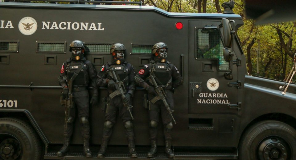 Agentes de segurança em trajes táticos ao lado de um veículo blindado durante uma operação.